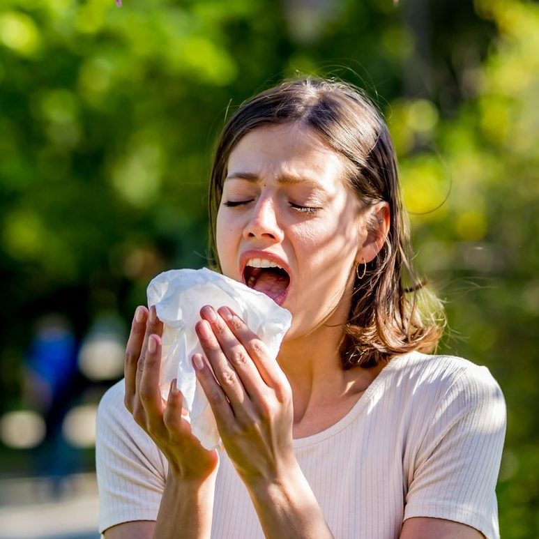 Una chica estornudando en plena calle al lado de la vegetación.