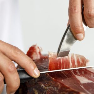 Hands of male butcher cutting Iberian ham at shop