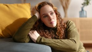 Portrait of a young woman with long curly red hair sitting on the floor and leaning on a sofa, looking away with sad face. Moment of sadness and worry in the living room of her house.