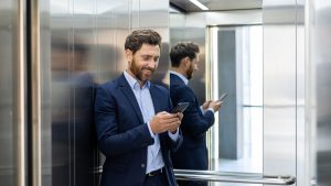 Smiling businessman in suit using smartphone in modern elevator, reflecting success and connectivity