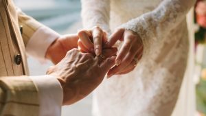 Bride putting ring on groom's finger. Rings exchange. Happy couple celebrating wedding outdoors in summer.