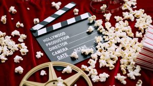 Clapperboard, film reel, film and spilled popcorn on red satin background.