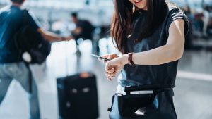 Cropped shot of young Asian woman with suitcase and passport, checking time on wristwatch in the airport concourse. Travel and vacation concept. Business person on business trip